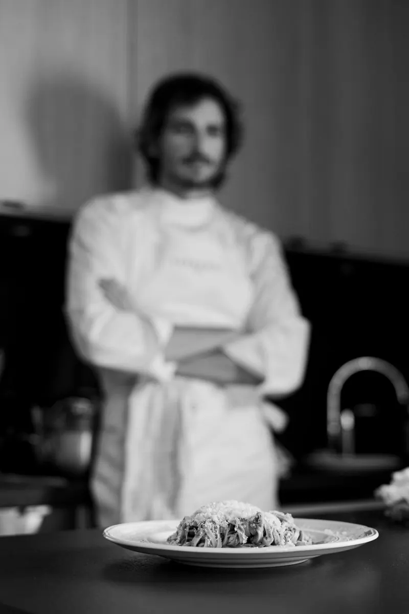 Assiette de tagliatelles au parmesan au premier plan, le chef flou en arrière-plan, en noir et blanc